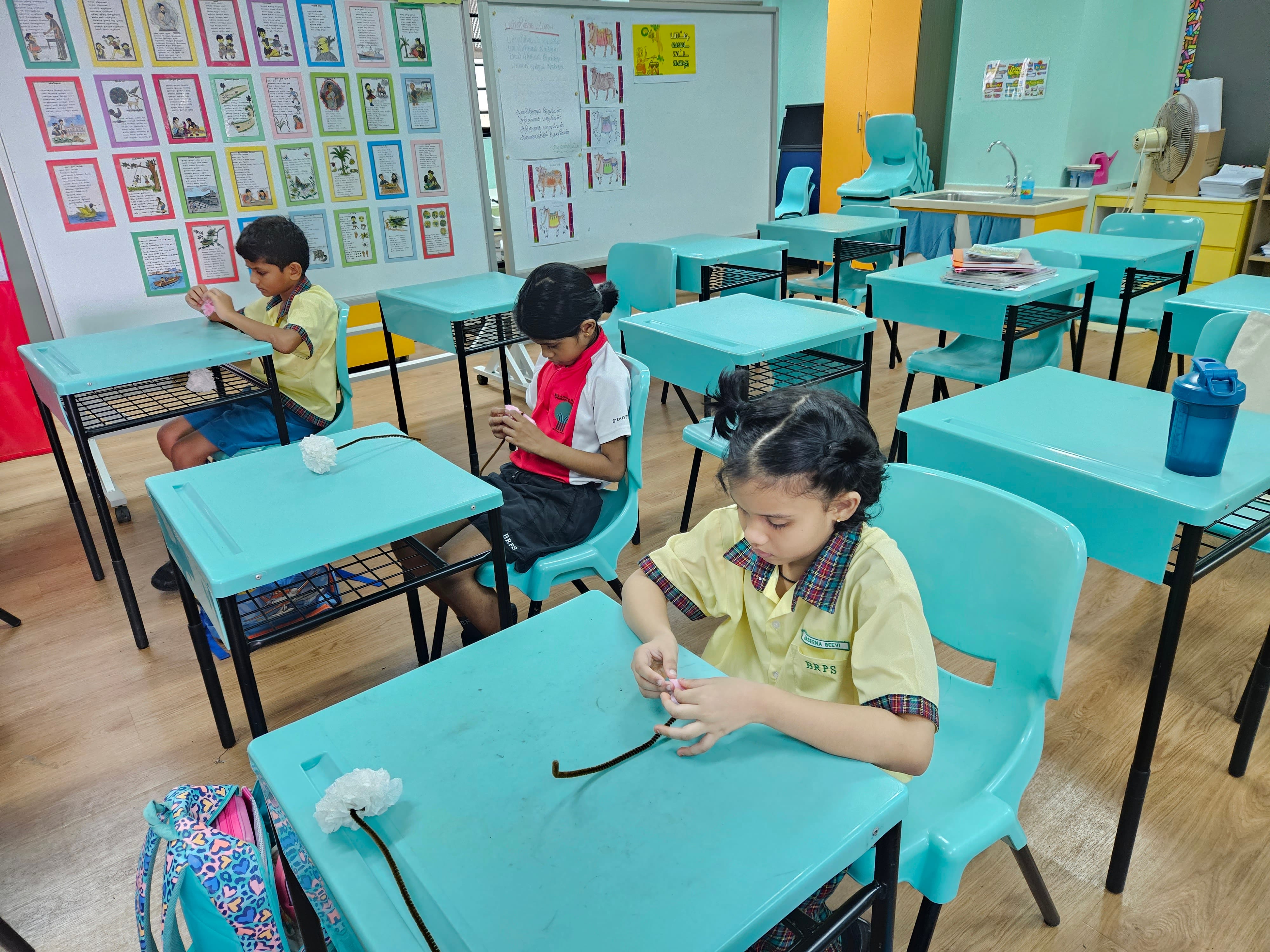 Three young students in uniforms sit at teal desks in a colorful classroom, working on a craft activity.